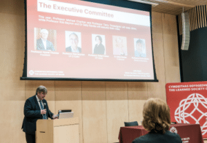 Professor Faron Moller standing at a lectern delivering a speech with a screen behind him displaying the names and faces of the LSW's governance members.