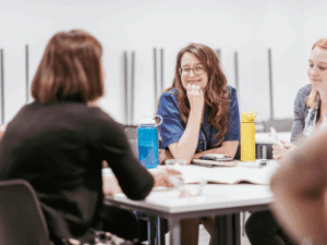A woman sits at a table smiling, with her hand on her chin, listening to another woman with her back to the camera. Papers and water bottles are on the table.