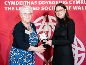Two women, one holding a Learned Society of Wales medal, shaking hands in fron of an LSW banner.