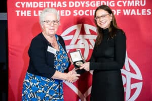 Two women, one holding a Learned Society of Wales medal, shaking hands in fron of an LSW banner.