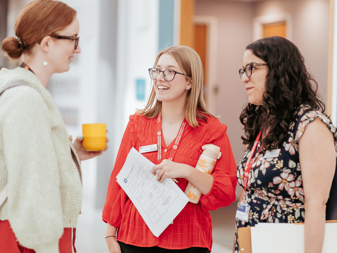 Three women, one holding a cup, the other two holding clipboards, in light conversation.
