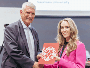 A woman and a man shaking hands while holding a brochure with the Learned Society of Wales logo on it.