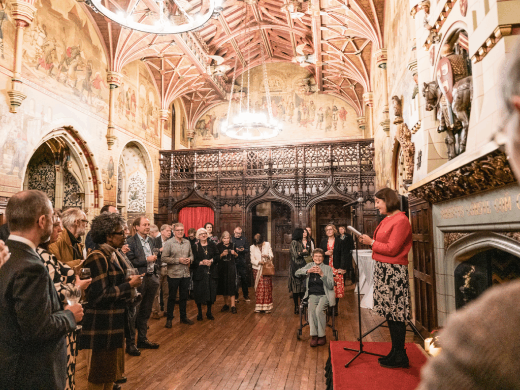 The inside of Cardiff Castle with ornate architecture and furnishings where a crowd of people are listening to a woman delivering a speech.