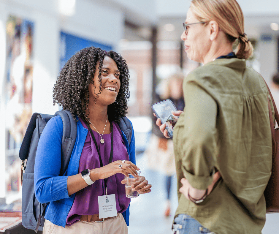 A woman, smiling, is talking to another woman who is holding a phone in her hand.