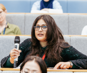 A woman, sitting in a lecture hall, holding a microphone.