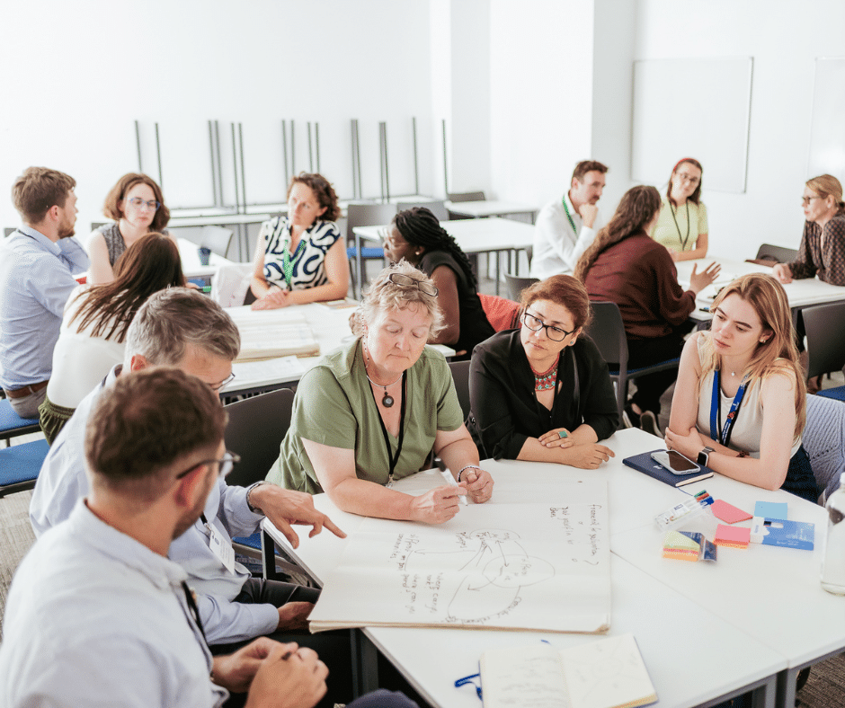 A group of people sit around a table with a large sheet of paper on which they are writing.