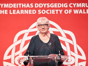 A woman at a lectern in front of a Learned Society of Wales banner stares straight at the camera.