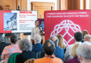 A woman at a lectern in from of a Learned Society of Wales banner talking to a room of people.